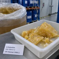 Yellow solid lumps of 4-Vinylaniline (4-Aminostyrene) CAS 1520-21-4 in a white plastic tray with a product label, beside an open fiber drum of lumps in a warehouse.