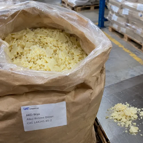 Open paper bag showing pale yellow AKD Wax Alkyl Ketene Dimer flakes CAS 144245-85-2 with a product label and flakes on a stainless steel table.