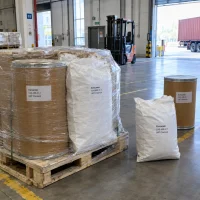 Pallet with shrink-wrapped fiber drums and white woven bags of Curcumin CAS 458-37-7 in a warehouse loading area with a forklift in the background.