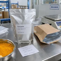 Silver foil bag of Curcumin CAS 458-37-7 next to a bowl of orange powder and a handwritten batch label on a stainless steel table in the packaging area.