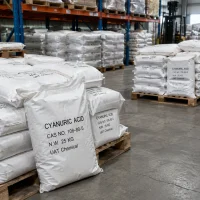Stacked 25kg white woven bags of Cyanuric Acid CAS 108-80-5 on wooden pallets in a warehouse with racks and a forklift in the background.